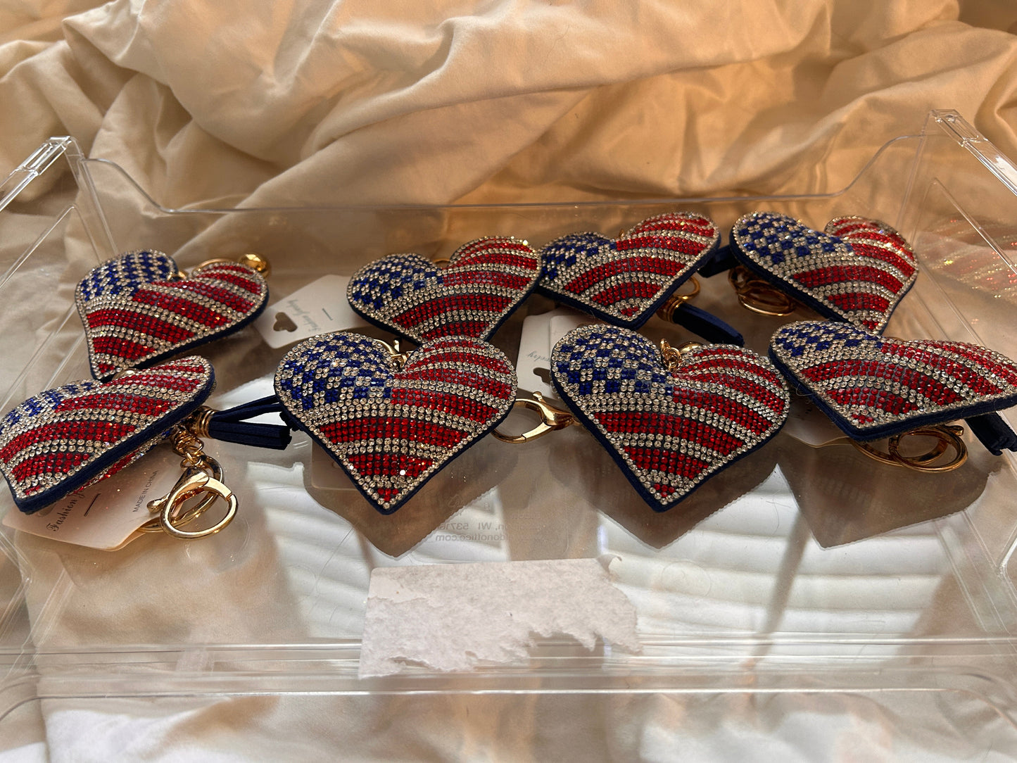 This Wrapables heart keychain features a puffy design adorned with red, white, and blue rhinestones forming an American flag pattern.It includes a gold-toned key ring, a lobster clasp for attachment to purses or handbags, and a dark blue tassel.The heart charm measures approximately 2.5 inches by 2.5 inches.The reverse side is made of a soft plush velvet material.