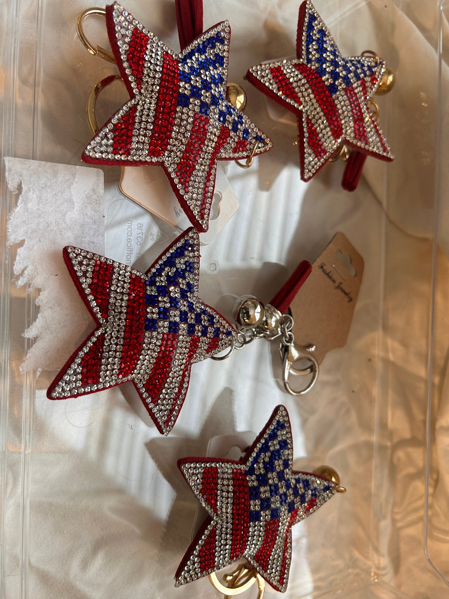 This Wrapables star keychain features a puffy design adorned with red, white, and blue rhinestones forming an American flag pattern.It includes a gold-toned key ring, a lobster clasp for attachment to purses or handbags, and a dark blue tassel.The star charm measures approximately 2.5 inches by 2.5 inches.The reverse side is made of a soft plush velvet material.