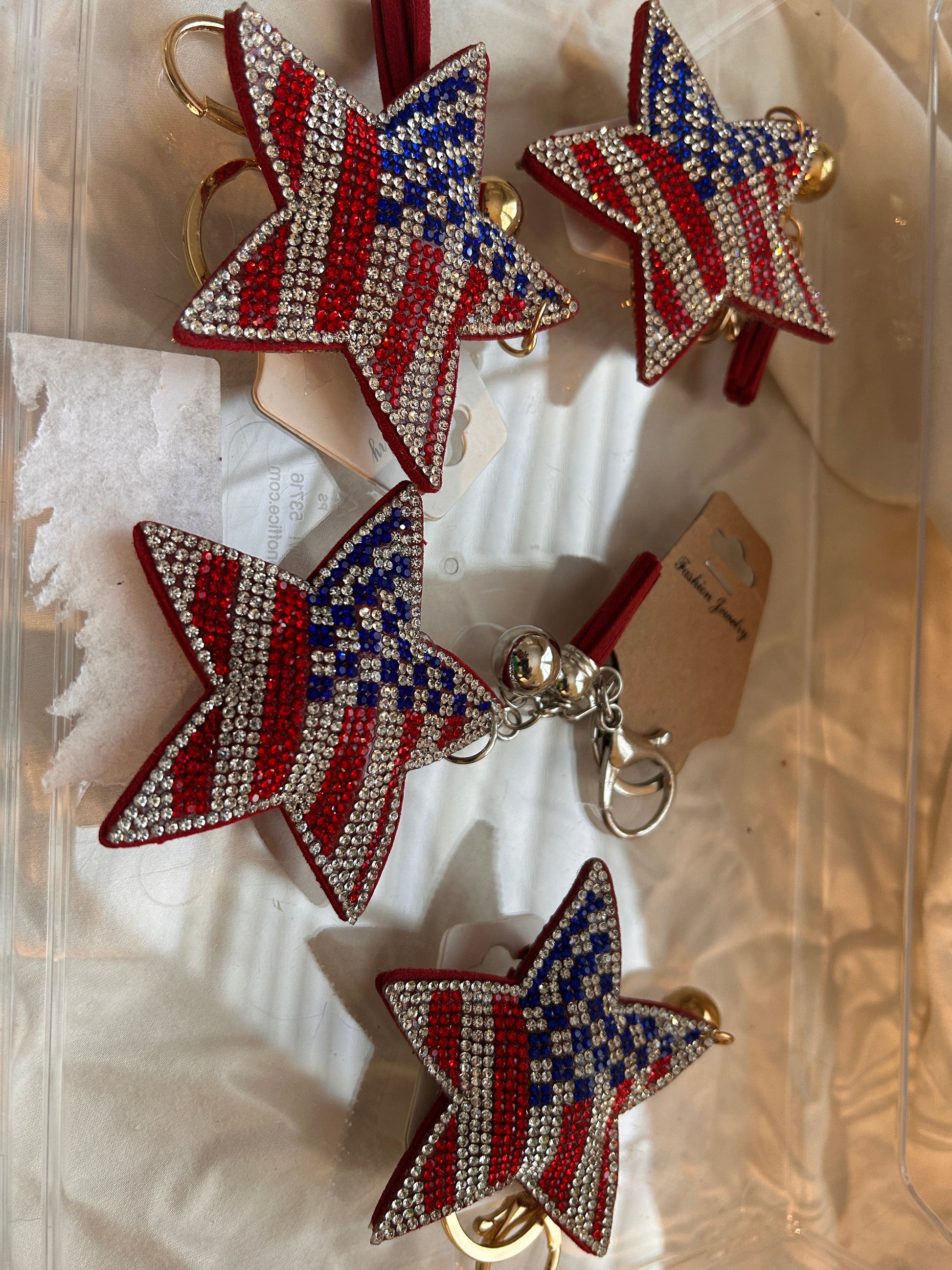This Wrapables star keychain features a puffy design adorned with red, white, and blue rhinestones forming an American flag pattern.It includes a gold-toned key ring, a lobster clasp for attachment to purses or handbags, and a dark blue tassel.The star charm measures approximately 2.5 inches by 2.5 inches.The reverse side is made of a soft plush velvet material.