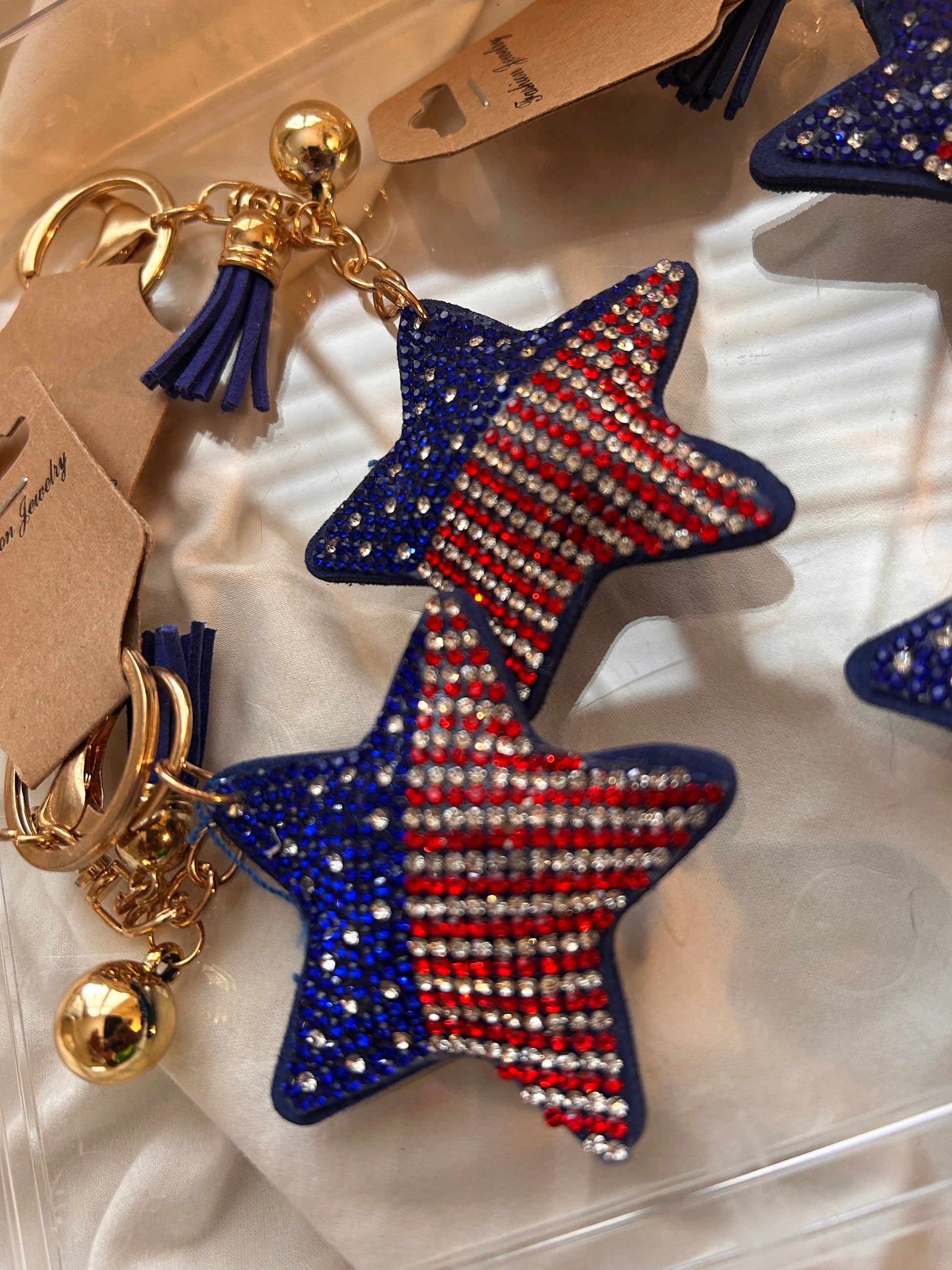 This Wrapables star keychain features a puffy design adorned with red, white, and blue rhinestones forming an American flag pattern.It includes a gold-toned key ring, a lobster clasp for attachment to purses or handbags, and a dark blue tassel.The star charm measures approximately 2.5 inches by 2.5 inches.The reverse side is made of a soft plush velvet material.