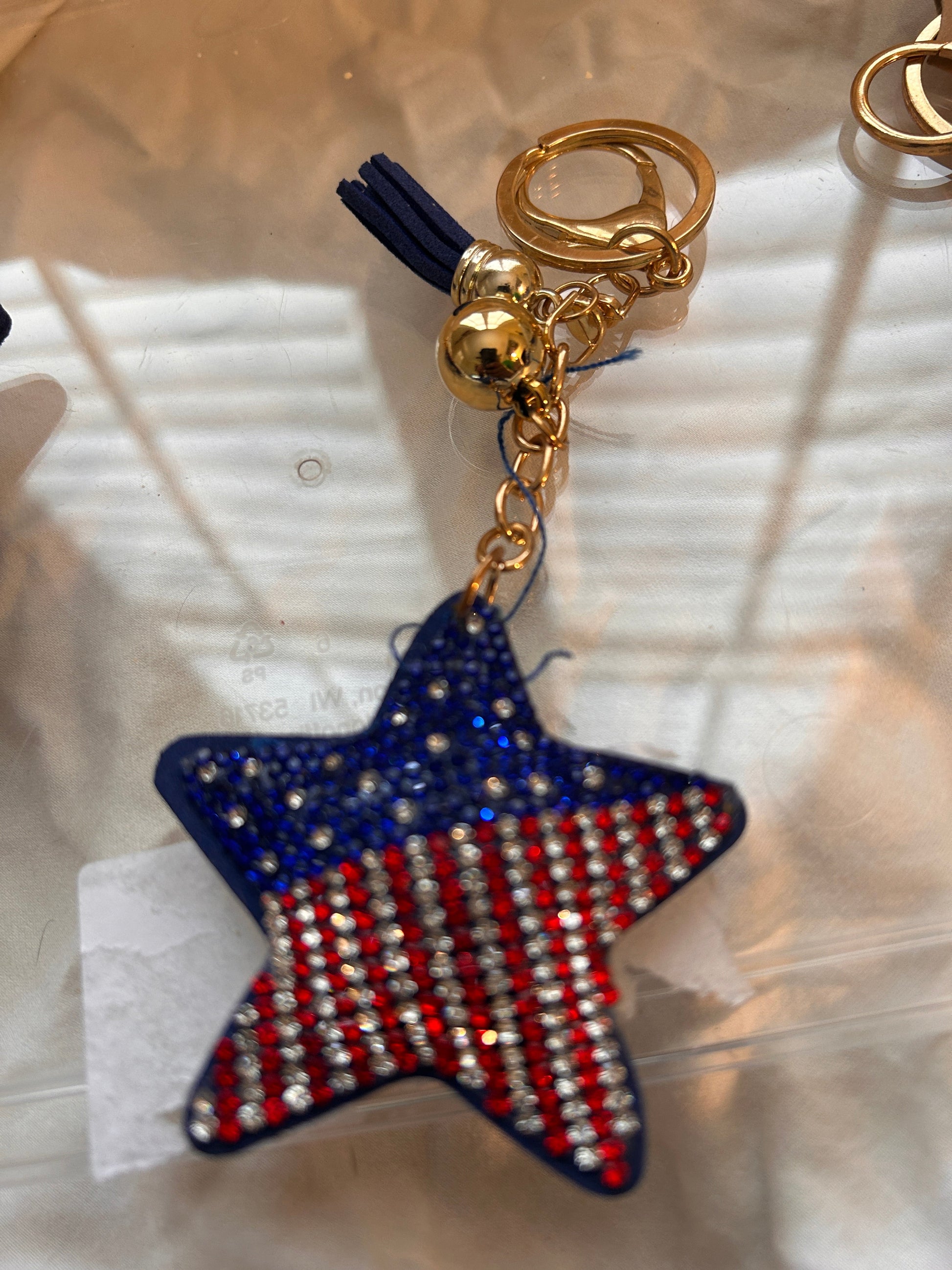 This Wrapables star keychain features a puffy design adorned with red, white, and blue rhinestones forming an American flag pattern.It includes a gold-toned key ring, a lobster clasp for attachment to purses or handbags, and a dark blue tassel.The star charm measures approximately 2.5 inches by 2.5 inches.The reverse side is made of a soft plush velvet material.