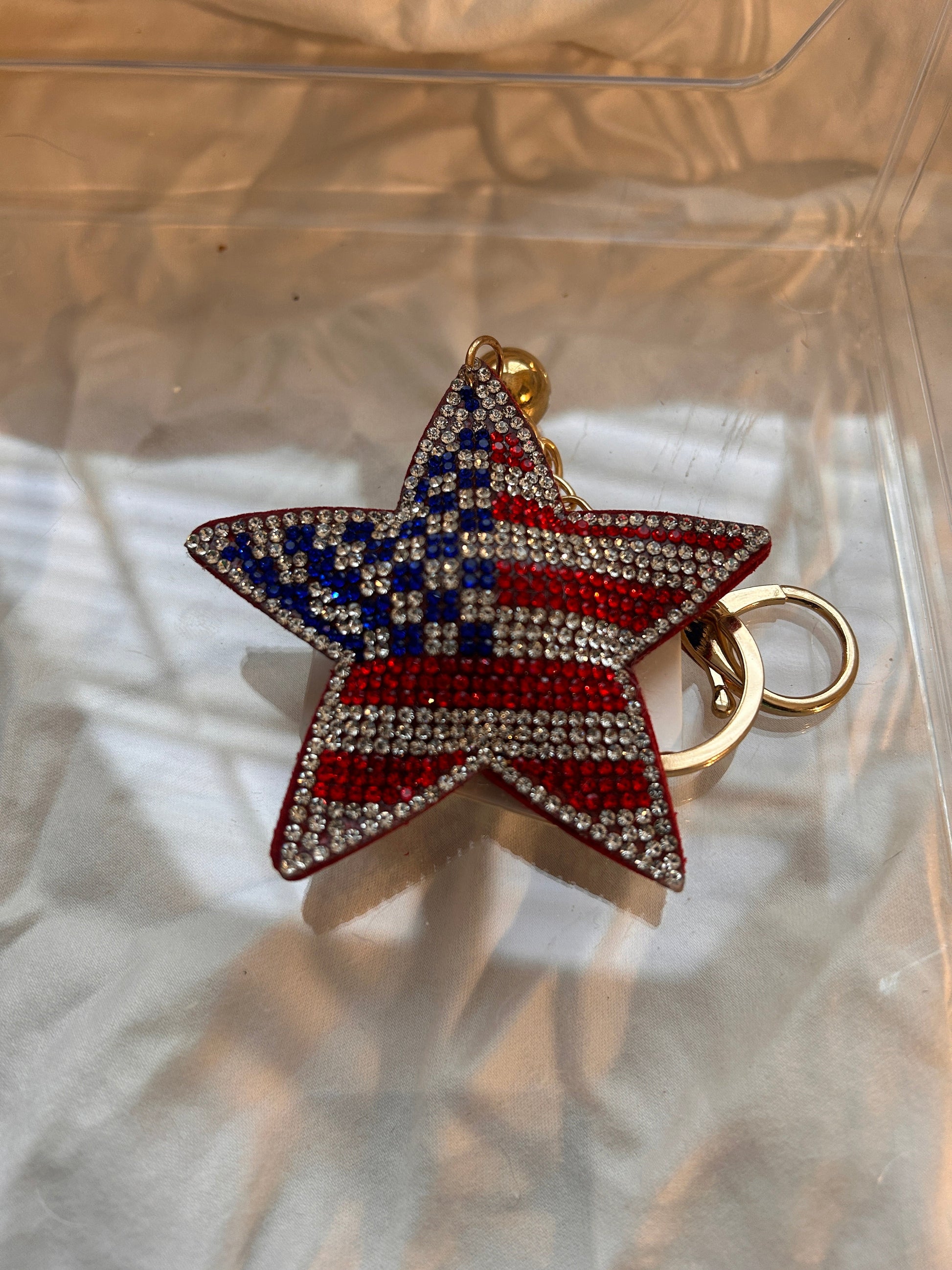 This Wrapables star keychain features a puffy design adorned with red, white, and blue rhinestones forming an American flag pattern.It includes a gold-toned key ring, a lobster clasp for attachment to purses or handbags, and a dark blue tassel.The star charm measures approximately 2.5 inches by 2.5 inches.The reverse side is made of a soft plush velvet material.